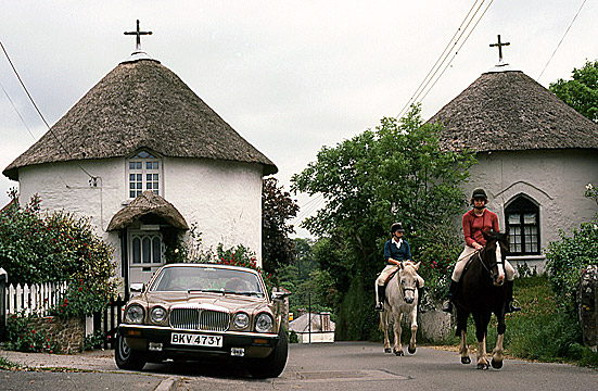 riders passing by thatched round houses and parked car in Veryan, Cornwall, southern England