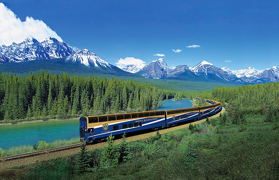 the Rocky Mountaineer making its way beside a stream with the Canadian Rockies in the background