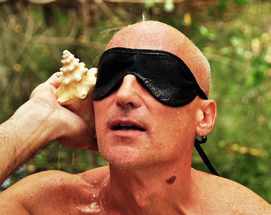 wet, blindfolded tourist holding a conch shell to his ear