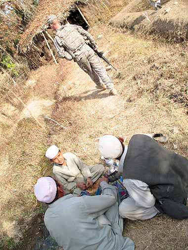 three Afghan kids sitting down for a meal as US Army soldier passes by