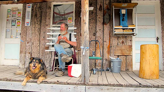 old man rolling a cigarette with a dog; pay-phone to his right, Atlanta, Idaho